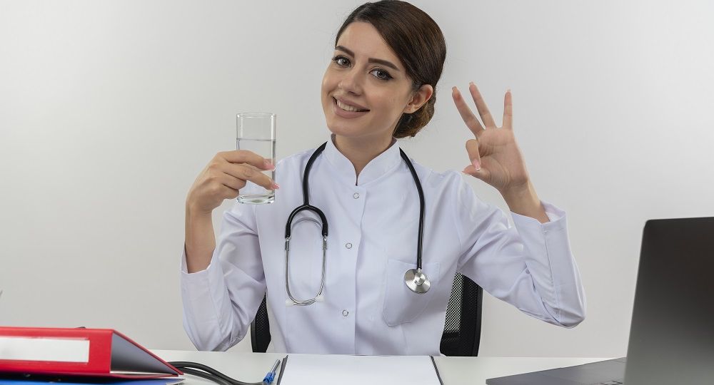 smiling young female doctor wearing medical robe with stethoscope sitting at desk work on computer with medical tools holding glass of water and showing okey gesture on isolated white background with copy space